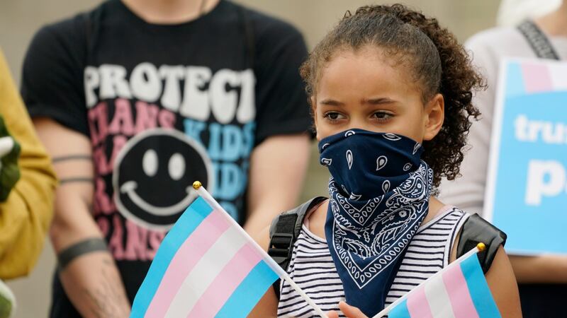 A young child holds a pair of trans pride flags at a noon gathering on the steps of the...